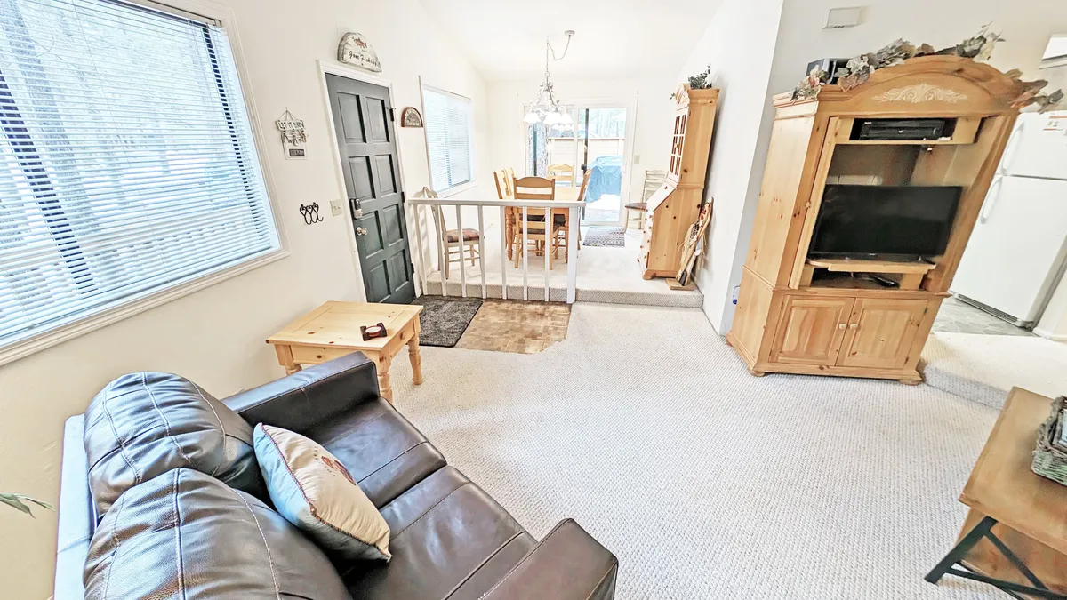 A bright and welcoming living/dining area, with a plush sofa, a spacious dining table, and entertainment unit. The space is designed for both relaxation and group meals, with décor that evokes the soothing tones of the forest. Ample natural light streams in, suggesting a connection to the outdoors.