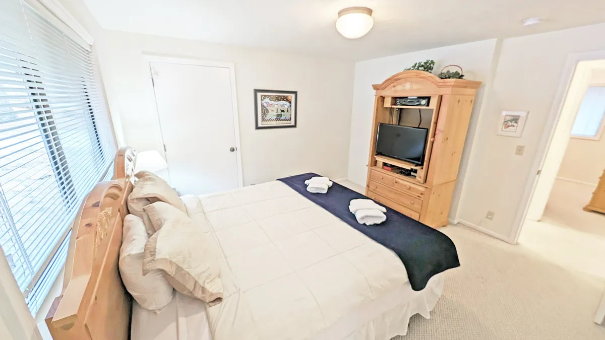 A tranquil bedroom scene, showcasing a comfortable bed with soft linens, an entertainment unit, and natural light filtering through window blinds. Tasteful wall art and soothing colors contribute to a restful, intimate atmosphere ideal for couples.