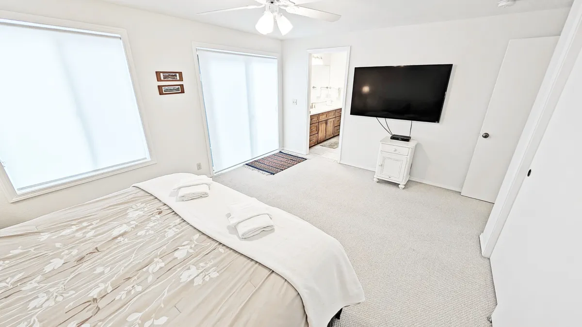 A spacious bedroom with a large king bed adorned with floral bedding sits beneath a wall-mounted TV. The room is brightened by multiple windows with sheer coverings, creating a soft, tranquil ambiance perfect for relaxation or catching up on a favorite show.