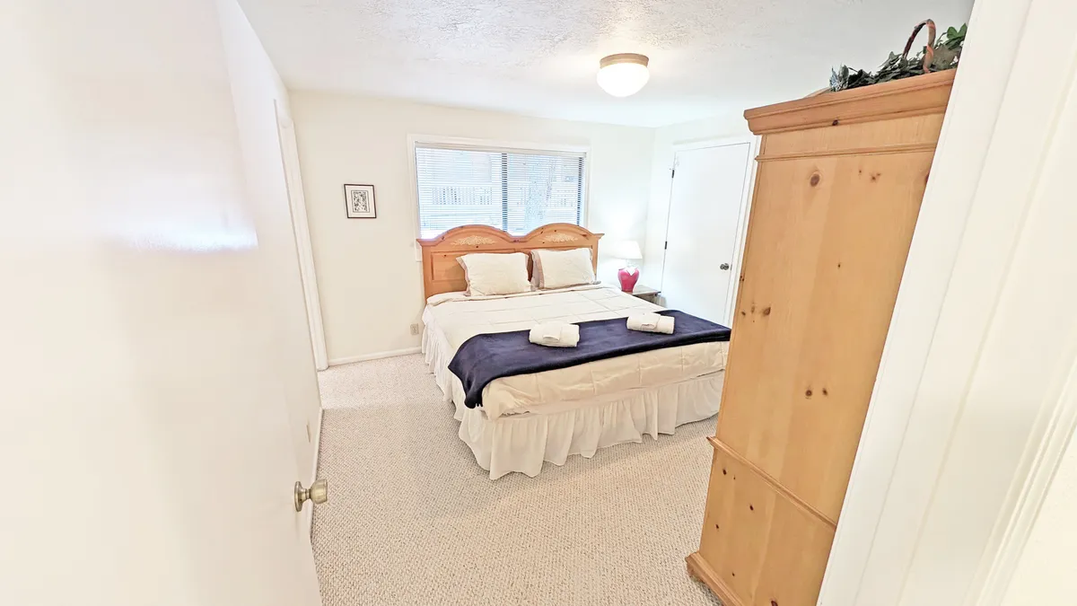This image features a serene bedroom with a king-sized bed dressed in a navy blanket and crisp white linens. Natural light streams in from a window, and a wooden wardrobe provides storage. The tranquil atmosphere and cozy details suggest a restful space perfect for unwinding.