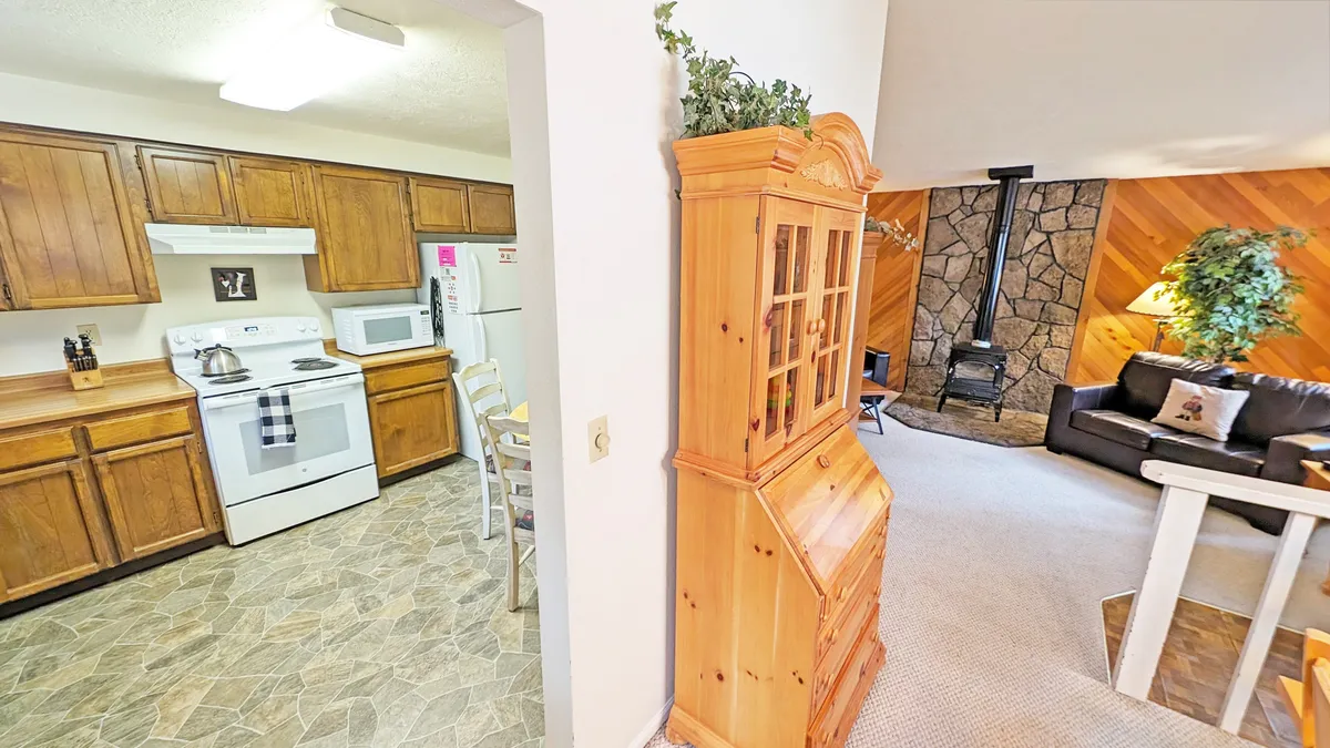 This image presents a welcoming open-concept space combining the kitchen and living room, featuring wooden furniture, comfortable seating, and a beautiful stone fireplace. The view extends from the kitchen to the cozy living area, creating a sense of flow and sociability. The décor subtly nods to the surrounding forest, enhancing the retreat’s rustic charm.