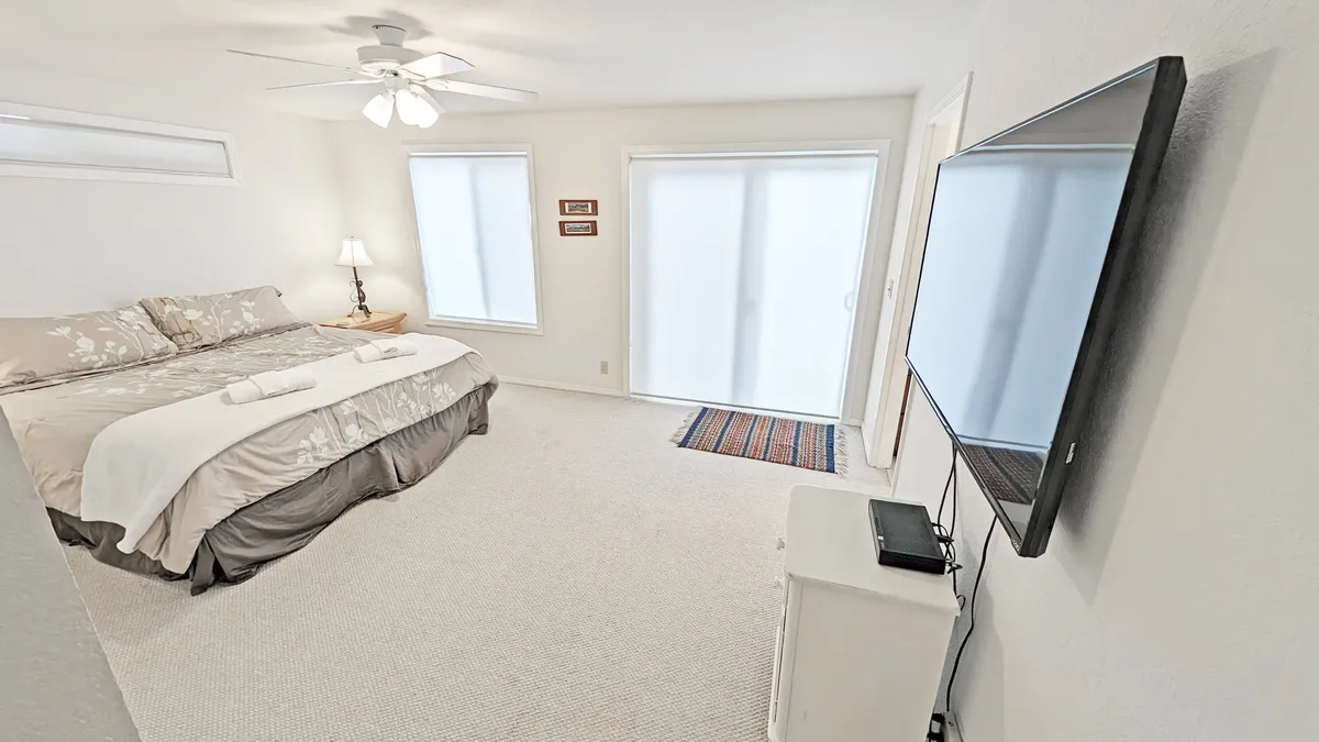 A serene bedroom featuring a bed with decorative pillows, a television, lamps, and sliding glass doors that open out to the surroundings. The room offers a sense of tranquility and direct access to the outdoors.