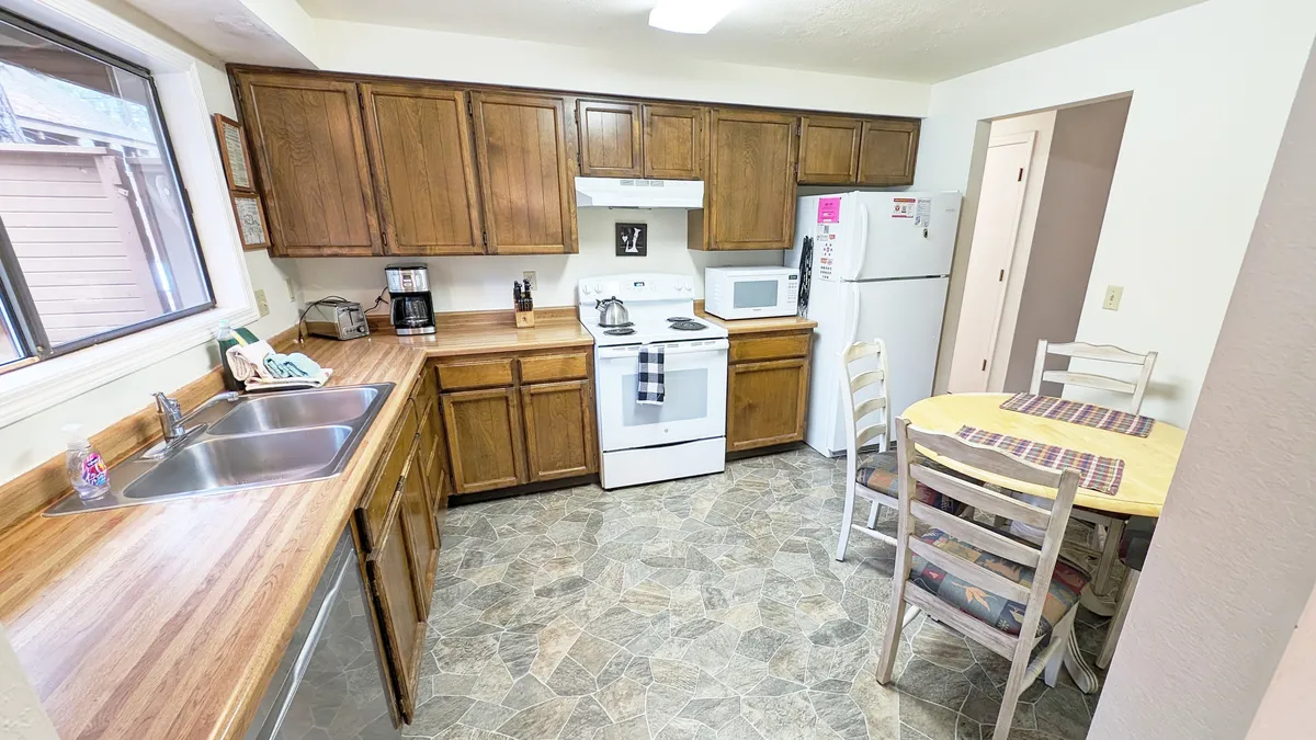 A view over a spacious kitchen area with wood cabinetry, a central stove, and a dining table set for meals or social gatherings. The scene suggests an open layout that connects seamlessly to the living space, ideal for shared activities and conversations.